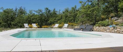 A rectangular swimming pool with a wooden deck in the foreground and lounge chairs around the pool.