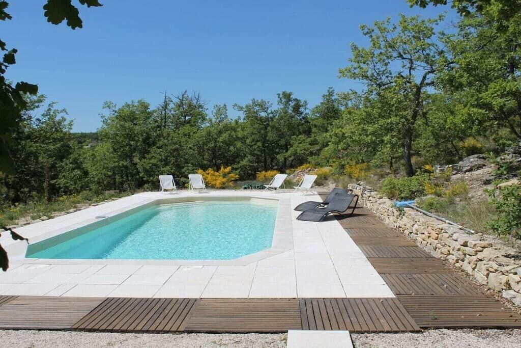 Une piscine rectangulaire entourée d’une terrasse en bois, avec des chaises longues blanches et un mur en pierre d’un côté.