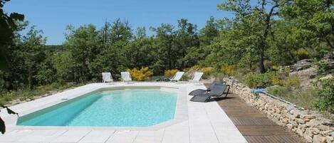 Une piscine rectangulaire entourée d’une terrasse en bois, avec des chaises longues blanches et un mur en pierre d’un côté.
