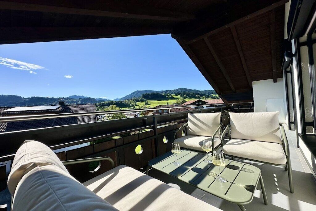 A balcony with a view of a green landscape and hills, a table with a glass of wine, and two cushioned chairs.