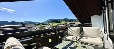 A balcony with a view of a green landscape and hills, a table with a glass of wine, and two cushioned chairs.