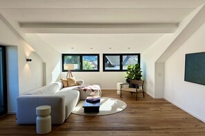 A modern living room with a sectional sofa, a coffee table, and a chair. There are windows allowing natural light and a view of the outdoors. - Holiday apartment with 1 bedroom (Wertach)