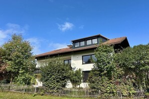 A two-story house with a sloped roof, surrounded by a wooden fence and lush greenery. - Holiday apartment with 1 bedroom (Wertach)