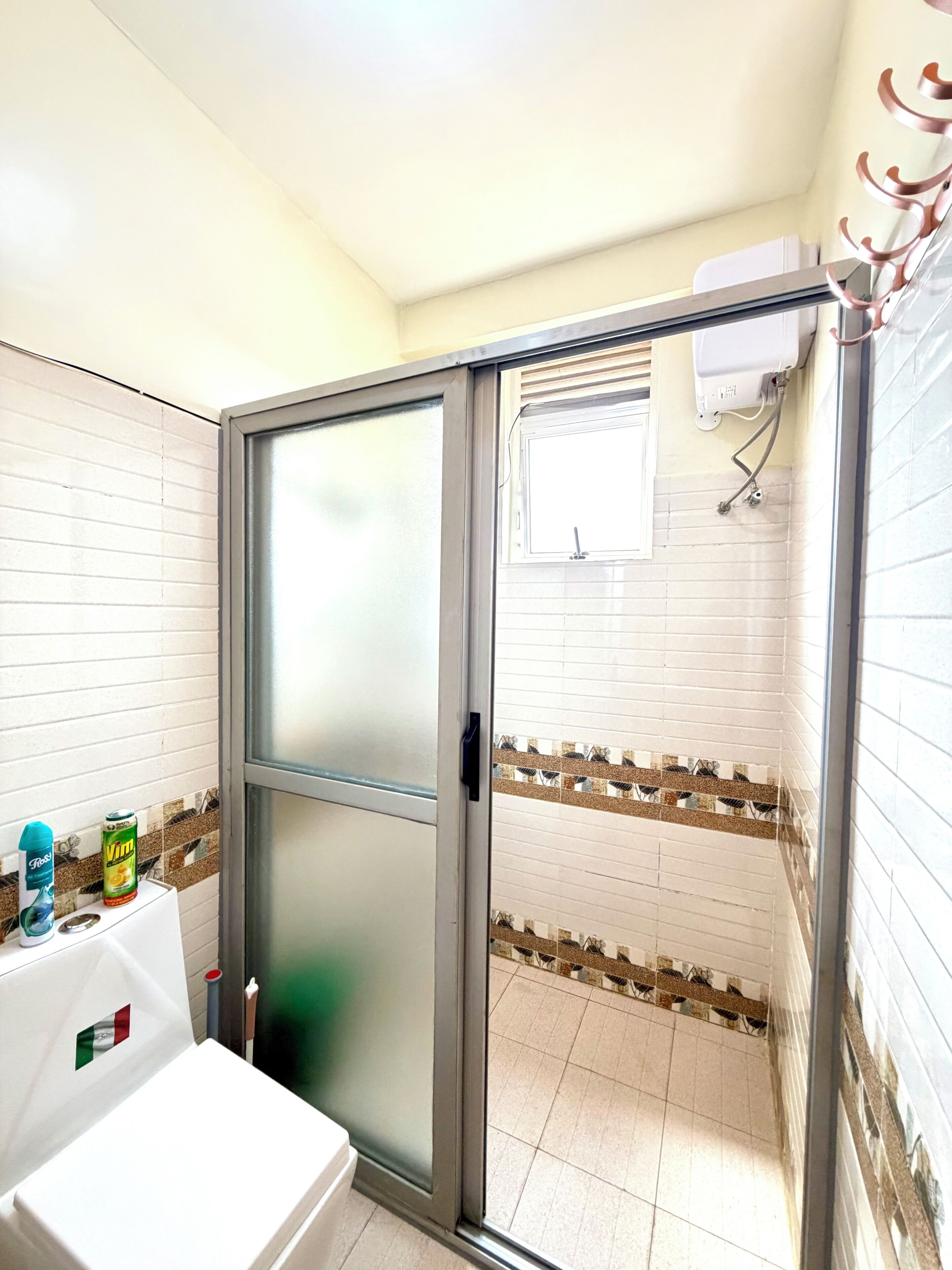Une salle de bain avec une douche à porte en verre, un lavabo décoré d’un motif à carreaux et une fenêtre où est accrochée une serviette.