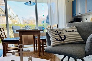 Ein modernes Wohnzimmer mit einem grauen Sofa, einem Holzesstisch und Blick ins Freie durch große Fenster.