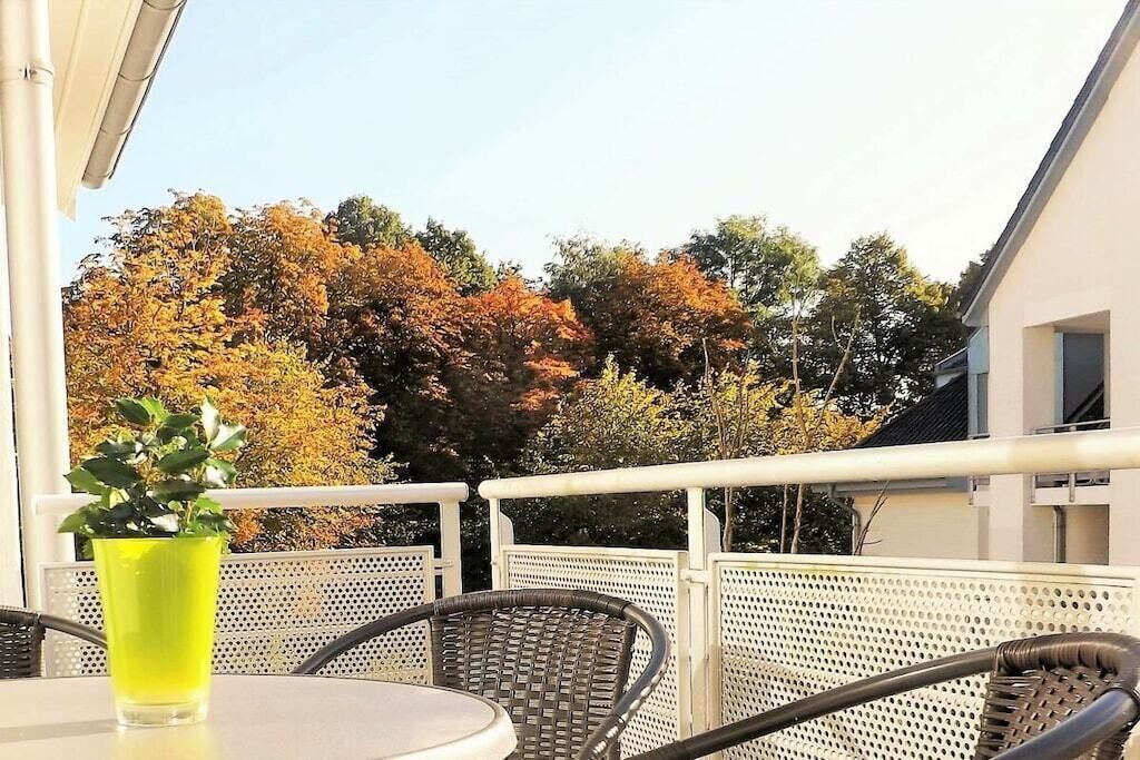 Een balkon met rieten stoelen en een tafel, een potplant en uitzicht op bomen met herfstbladeren.