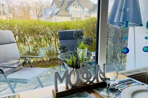 Une table en verre avec une plaque nominative, un verre et une chaise, dans une pièce offrant une vue sur des maisons et de la verdure.