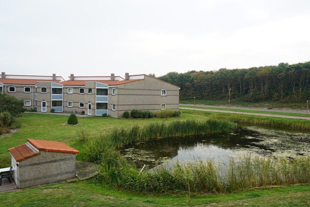 Un edificio residenziale su più piani, con un piccolo stagno e una zona erbosa alberata sullo sfondo.