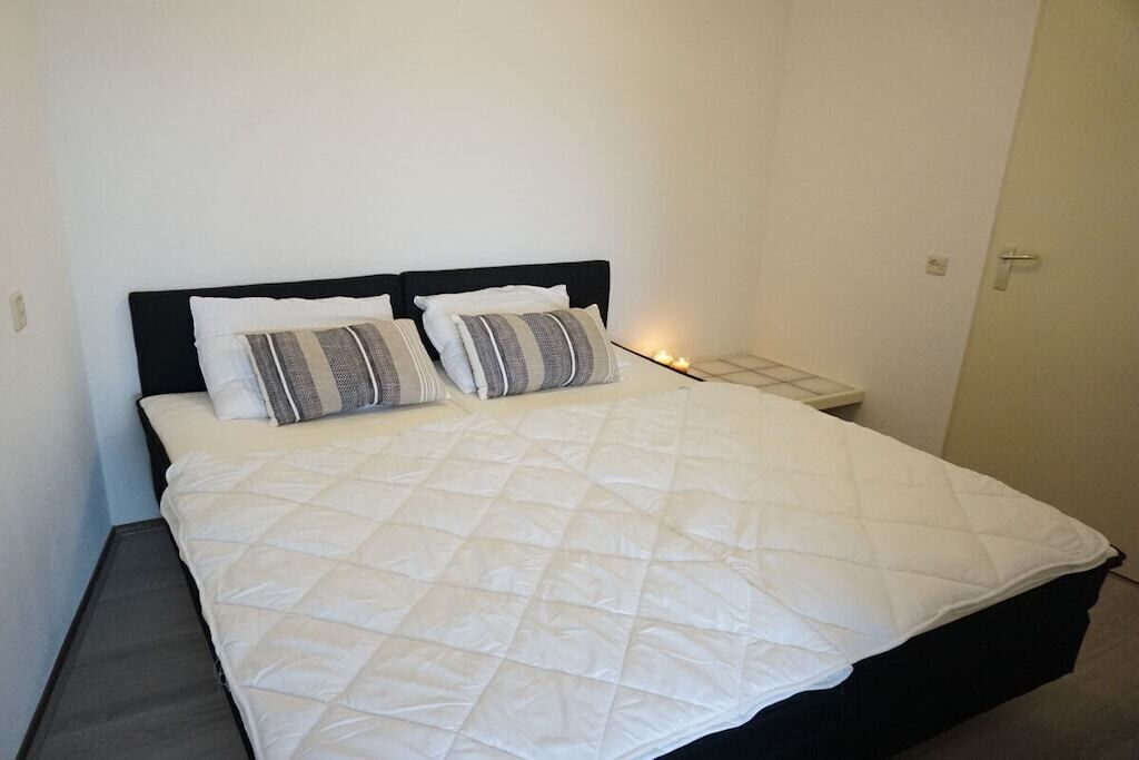 Un lit recouvert d’une courtepointe blanche et orné de deux oreillers, une table de chevet avec une lampe, et une porte sur la droite.