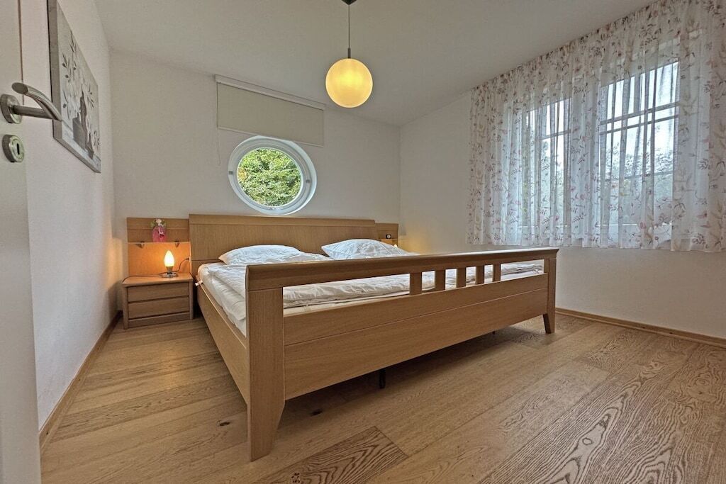 A bedroom with a wooden bed, a bedside table with a lamp, a round window, and a view of greenery outside.