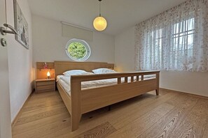 Une chambre avec un lit en bois, une table de chevet avec une lampe, une fenêtre ronde et une vue sur la verdure à l’extérieur.