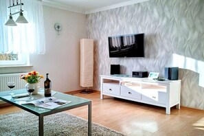 A modern living room with a flat-screen TV mounted on a textured wall, a white entertainment unit with a glass top, and a glass coffee table with a magazine and wine bottle. - Holiday apartment with 1 bedroom (Kühlungsborn Ost)