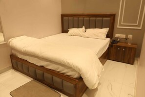 A neatly made bed with a wooden headboard and a small bedside table with a phone and a bottle. - Aachman Hotal Restaurant (Bulandshahr)