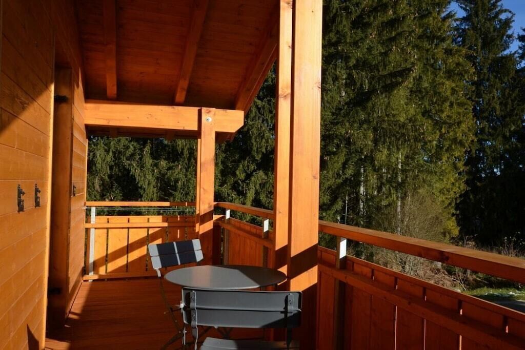 Une terrasse en bois avec une table et des chaises, entourée d’une balustrade en bois, et une vue sur les arbres en arrière-plan.