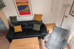 A living room with a black leather sofa, two wooden side tables, a grey armchair, and a colorful abstract painting on the wall. - Jardin de La Clape No. 21 (Narbonne-Plage)