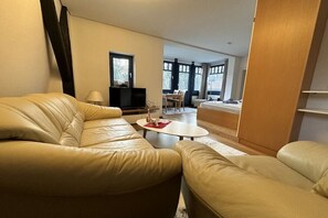 Ein Wohnzimmer mit einer Couch, einem Couchtisch und einem Fernseher. Es gibt einen Essbereich mit Tisch und Stühlen.