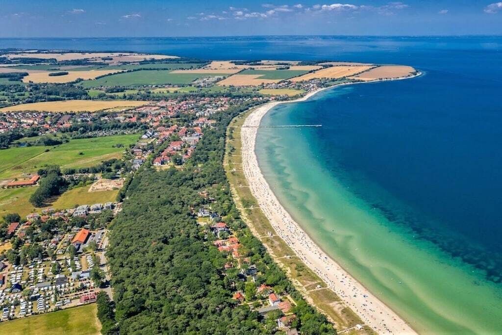 Luchtbeeld van een kustgebied met een lang zandstrand, een dichtbebouwd woongebied met huizen en een heldere blauwe zee.