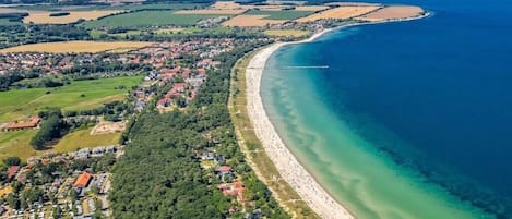 Eine Luftaufnahme einer Küstenregion mit einem langen Sandstrand, einem dicht bebauten Wohngebiet mit Häusern und einem klaren, blauen Meer.