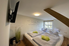 Un dormitorio con cama, almohadas, un televisor montado en la pared, una ventana y una pared decorativa con texto.