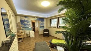 A room with a beach mural, a sofa, a chair, a plant, and a window with blinds. - Holiday home "On the cliffs" Apt. 3 (Boltenhagen Redewisch)