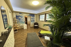 Una habitación con un mural de playa, un sofá, una silla, una planta y una ventana con persianas.
