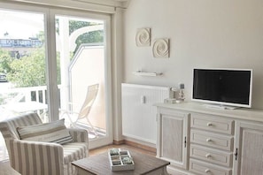 A living room with a TV, white furniture, a window, and a view of greenery outside. - Apartment Residence Baltic Sea Beach (Kühlungsborn)