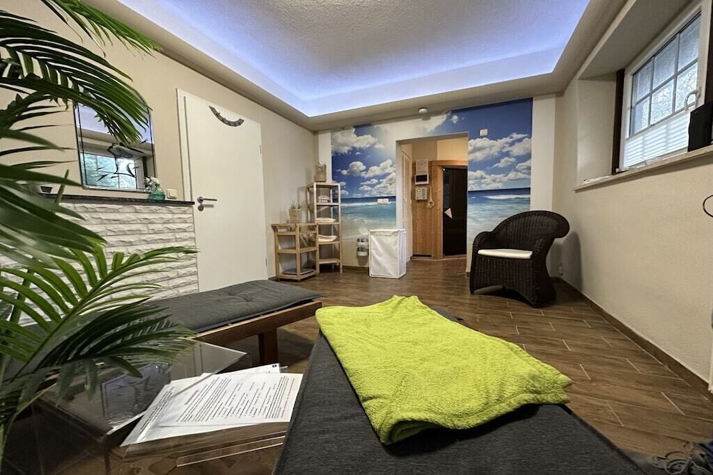 Ein Zimmer mit einem Bett, einem Sessel, einem kleinen Tisch, einer Pflanze und einem Meeresgemälde.