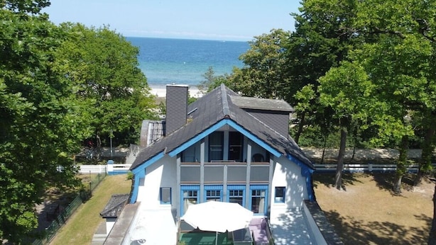 Una casa de dos pisos con fachada azul y blanca, un porche con un banco verde y vista al océano.