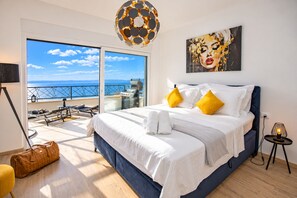 Ein Hotelzimmer mit einem großen Bett, Meerblick, einem Balkon mit Grill und einem modernen Kronleuchter.