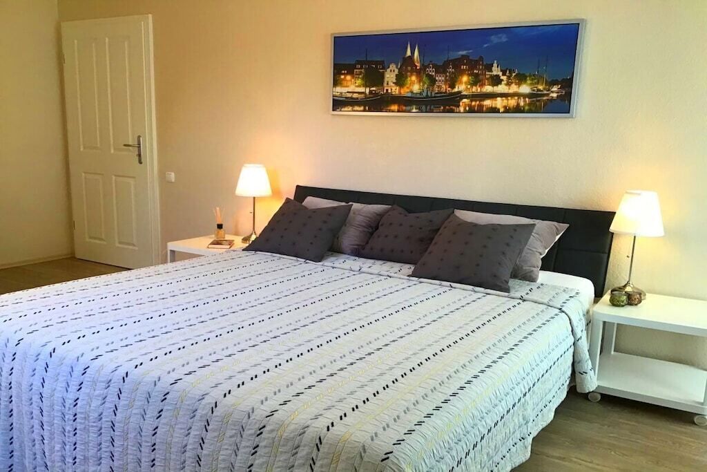 Ein ordentlich bezogenes Bett mit einem gemusterten Überwurf, zwei Nachttischlampen und einem gerahmten Stadtbild an der Wand.