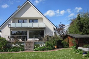 Una casa moderna de dos pisos con un balcón, un pequeño jardín y un cobertizo de madera.