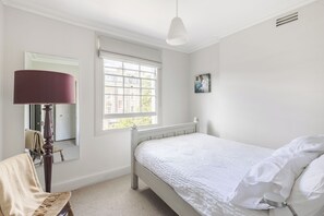 Ein Schlafzimmer mit Bett, Fenster, Lampe, Spiegel und einem Gemälde.