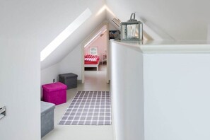 Ein schmales Dachzimmer mit schrägem Dach, einem Schachbrettteppich und einem Bett mit einer rot-weiß karierten Bettdecke.