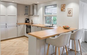 Une cuisine moderne avec un plan de travail en bois, un four encastré et une fenêtre donnant sur un paysage verdoyant. On y trouve deux tabourets de bar et un saladier rempli de fruits sur le plan de travail.