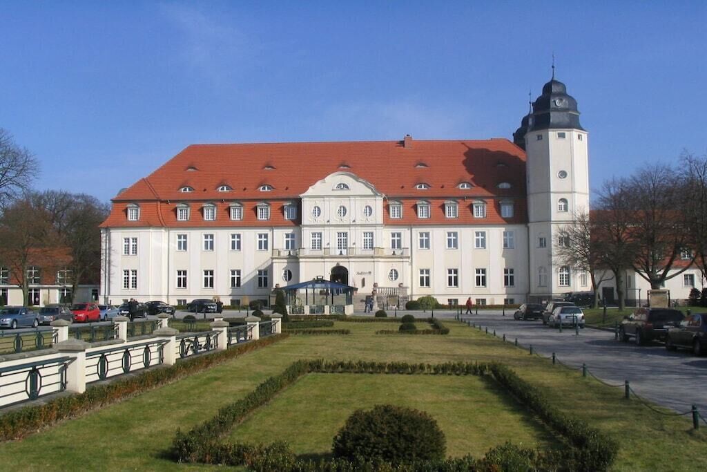 A large, historic building with a central tower, a symmetrical facade, and a well-maintained garden in front.