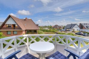 En balkong med ett vitt bord och blå stolar, med utsikt över ett bostadsområde med hus och parkerade bilar.