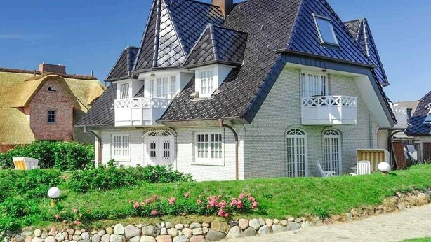 Une maison à deux étages, dotée d’un toit à forte pente, de nombreuses fenêtres et d’un mur de pierre en premier plan.