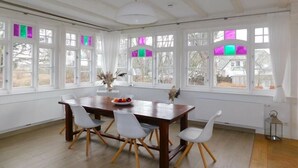 Uma sala de jantar com mesa de madeira e cadeiras brancas, janelas amplas com cortinas e vista para o exterior.