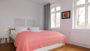 Quarto com cama, mesa de cabeceira, janela com cortinas e radiador.
