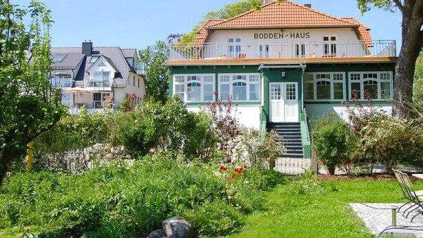 Um edifício de dois andares chamado 'Bodden-Haus' com uma varanda, rodeado por vegetação e um jardim.