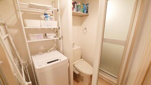 A compact bathroom with a white toilet, a small shelf with toiletries, and a white washing machine. - Sakura Court Ebisu (Shibuya)