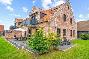 Une maison à deux étages, construite en briques, dotée d’un balcon, d’un espace extérieur aménagé pour s’asseoir et d’un petit jardin.