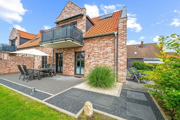 Une maison à deux étages, construite en briques, dotée d’un balcon, d’une terrasse avec une table et des chaises, et d’un jardin.