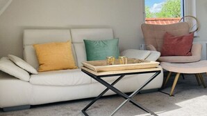 A living room with a white sofa, a small round table, and a window with a view of trees. - Holiday apartment in Nesse with swimming pool (Nesse)
