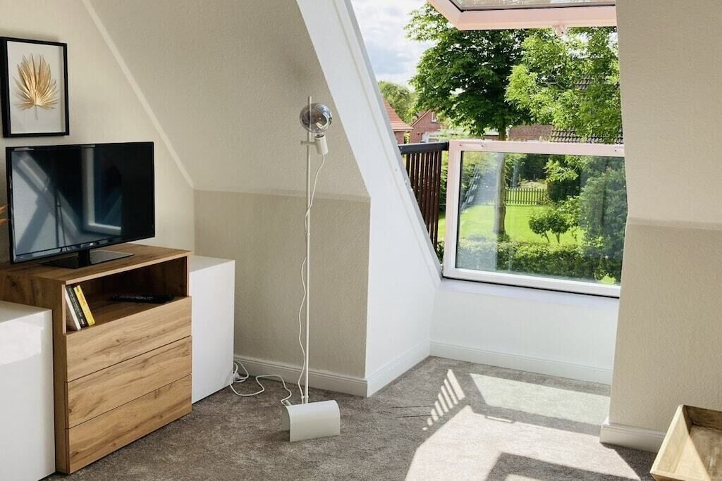 Ein Zimmer mit Fernseher auf einem Holzboden, einer Stehlampe und Blick auf eine grüne Landschaft durch ein Fenster.