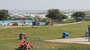 A grassy area with a playground, including swings and a slide, and a pool area with umbrellas and lounge chairs. - Holiday apartment in Nesse with swimming pool (Nesse)