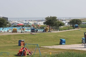Una zona verde con un'area giochi, con altalene e uno scivolo, e una piscina con ombrelloni e lettini.