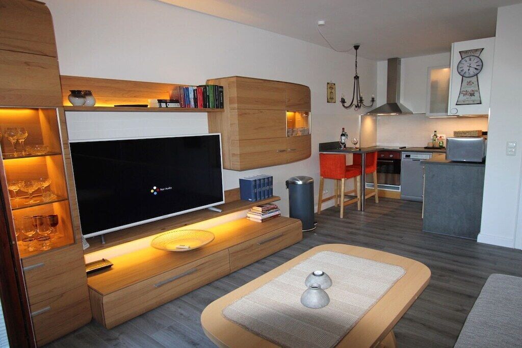 Ein modernes Wohnzimmer mit Flachbildfernseher, Holzschränken, einer Essecke mit Tisch sowie einer Küche mit Mikrowelle und Herd.