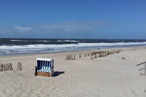 Ein Strand mit einem einzelnen gestreiften Liegestuhl und einer Holzkonstruktion, möglicherweise einer Rettungswache, mit dem Meer im Hintergrund.