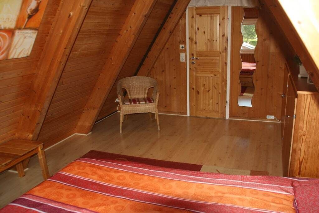 A cozy attic bedroom with a wooden sloped ceiling, a single wicker chair, a bed with a patterned bedspread, and a wooden door leading to another room.